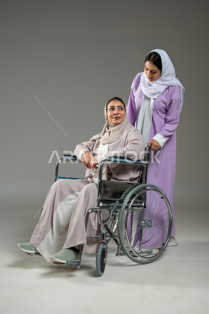 العنايه بأصحاب الهمم، شعور المعاقين بالإنتماء والتقدير، بورتريه لشابة عربية سعودية خليجية محجبة تردي العباءة تقوم بمساعدة امرأة من ذوي الاحتياجات الخاصة، تأمين اجهزة ومعدات تساعد بالحركة والتنقل، الجسم بالطول الكامل، خلفية رمادية، محتوى حساس، استخدام حساس