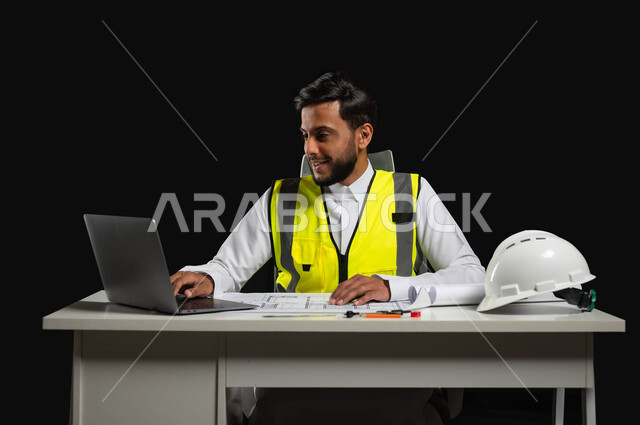 Looking at the mobile device with an expression of pleasure, a close-up portrait of a Saudi Gulf Arab engineer wearing a traditional dress and a work jacket sitting at a desk with architectural engineering plans in front of him, black background, sensitive use, sensitive content