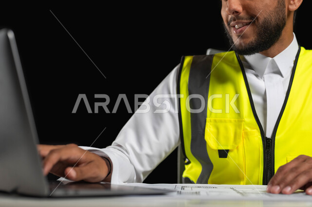 Studying the basics of the project through laptop applications, a close-up portrait of a Saudi Gulf Arab engineer wearing a traditional dress and a work jacket sitting at a desk with architectural engineering plans in front of him, black background, sensitive use, sensitive content