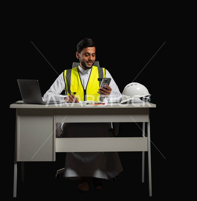اعتماد التقنيات الحديثة في الهندسة, دمج البيئة العملية و الهندسه مع  التكنولوجيا، بورتريه  لمهندس عربي خليجي سعودي يرتدي الثوب التقليدي و سترة الحماية يجلس وراء المكتب وينظر إلى شاشة الجوال, الجسم بالطول الكامل، خلفية سوداء، محتوى حساس، استخدام حساس