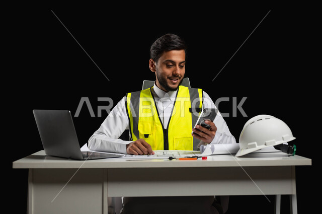 Integrating the practical environment and engineering with technology and technology, a close-up portrait of a Saudi Arabian Gulf engineer wearing a traditional dress and a protective jacket, sitting behind the desk and looking at the mobile screen with gestures of pleasure, building and construction engineering, black background, sensitive content, sensitive use