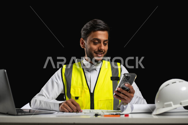 Using modern technologies and devices, browsing engineering applications and websites, a close-up portrait of a Saudi Arabian Gulf engineer wearing a traditional dress and a protective jacket, sitting behind the desk and looking at the mobile screen with happy gestures, building and construction engineering, black background, sensitive content, sensitive use