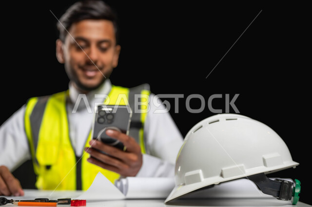 Working in the engineering field, browsing engineering applications and websites, a close-up portrait of a Saudi Arabian Gulf engineer wearing a traditional dress and a protective jacket, sitting behind the desk and looking at the mobile screen with happy gestures, building and construction engineering, black background, sensitive content, sensitive use