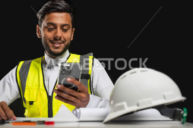 تصفح التطبيقات والمواقع الهندسية,  إدارة المشاريع الهندسية, بورتريه مقرب لمهندس عربي خليجي سعودي يرتدي الثوب التقليدي و سترة الحماية  يجلس وراء المكتب وينظر إلى شاشة الجوال بإيماءات السرور, هندسة الأبنية و الإنشاءات، , خلفية سوداء، محتوى حساس، استخدام حساس