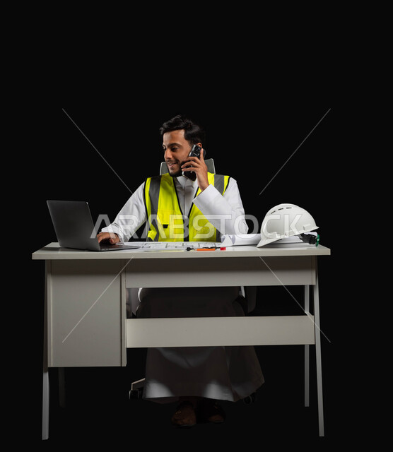 Completing work projects by making a phone call using a mobile phone, portrait of a Saudi Arabian Gulf engineer wearing a traditional dress and a protective jacket, sitting behind the desk and looking at the mobile device screen with pleased gestures, full-length body, black background, sensitive content, sensitive use