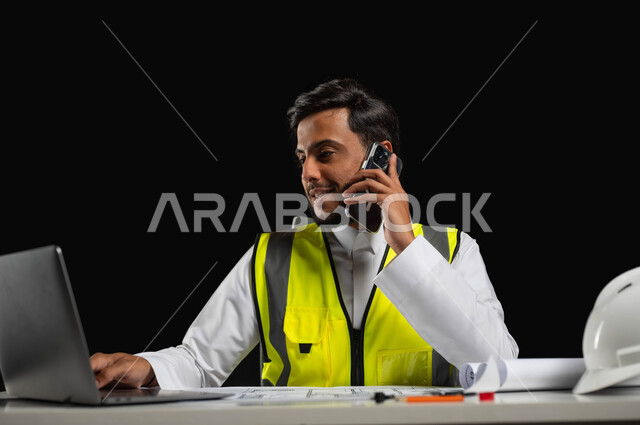 التواصل والدردشة مع الأصدقاء باستعمال الهاتف، استخدام التقنيات والأجهزة الحديثة, بورتريه مقرب لمهندس عربي خليجي سعودي يرتدي الثوب التقليدي و سترة الحماية  يجلس وراء المكتب وينظر إلى شاشة الجهاز المحمول بإيماءات السرور, خلفية سوداء، محتوى حساس، استخدام حساس