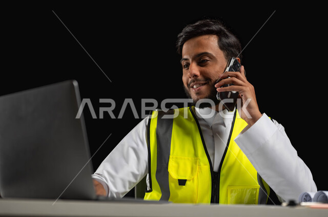 الرد على اتصالات الهاتف المحمول، استخدام التقنيات والأجهزة الحديثة, بورتريه مقرب لمهندس عربي خليجي سعودي يرتدي الثوب التقليدي و سترة الحماية  يجلس وراء المكتب وينظر إلى شاشة الجهاز المحمول بإيماءات السرور, خلفية سوداء، محتوى حساس، استخدام حساس