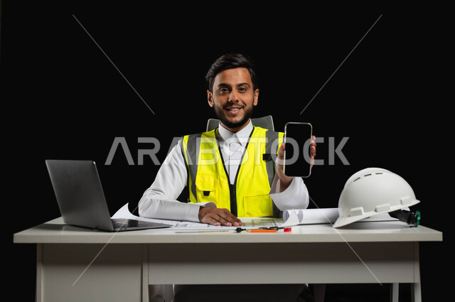 النظر إلى الكاميرا بإيماءات السرور، عرض شاشة سوداء فارغة،  دمج التقنية والتكنولوجيا في مجال الهندسة،  بورتريه مقرب لمهندس عربي خليجي سعودي يرتدي الثوب التقليدي مع سترة الحماية يجلس خلف المكتب، خلفية سوداء، استخدام حساس، محتوى حساس
