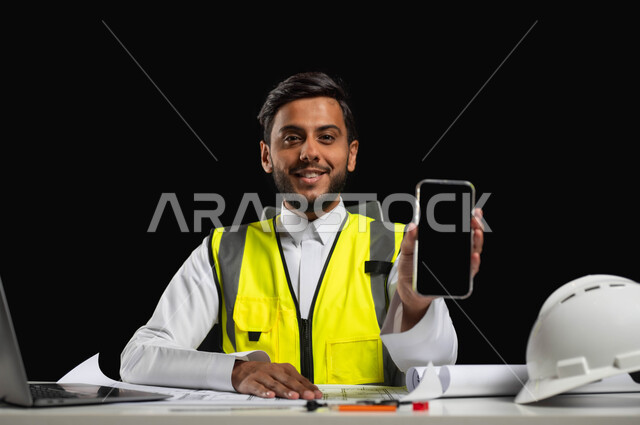 عرض شاشة سوداء فارغة، النظر إلى الكاميرا بإيماءات السرور، دمج التقنية والتكنولوجيا في مجال الهندسة،  بورتريه مقرب لمهندس عربي خليجي سعودي يرتدي الثوب التقليدي مع سترة الحماية يجلس خلف المكتب، خلفية سوداء، استخدام حساس، محتوى حساس