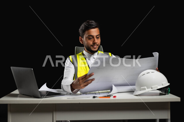 The concept of project management and auditing, developing and developing engineering fields, a close-up portrait of a Saudi Arabian Gulf engineer wearing the traditional dress and work jacket, sitting at the desk and holding engineering plans in his hand, supervising the work, black background, sensitive content, sensitive use