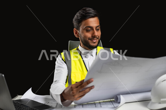 مفهوم الهندسة والإنشاءات المعمارية، بورتريه مقرب لمهندس عربي خليجي سعودي يرتدي الثوب التقليدي والسترة الخاصة بالعمل ويجلس على المكتب ويمسك بيده مخطط هندسي، إيماءات الفخر بالوصول إلى حلم النجاح، الإشراف على العمل، خلفية سوداء، محتوى حساس، استخدام حساس