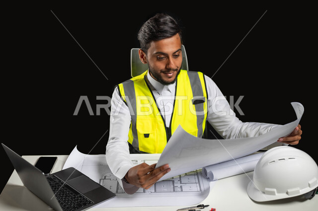 تنمية وتطوير المجالات الهندسية، مفهوم الهندسة والإنشاءات المعمارية، بورتريه مقرب لمهندس عربي خليجي سعودي يرتدي الثوب التقليدي و السترة الخاصة بالعمل ويجلس على المكتب ويمسك بيده مخططات هندسية، الإشراف على العمل، خلفية سوداء، محتوى حساس، استخدام حساس