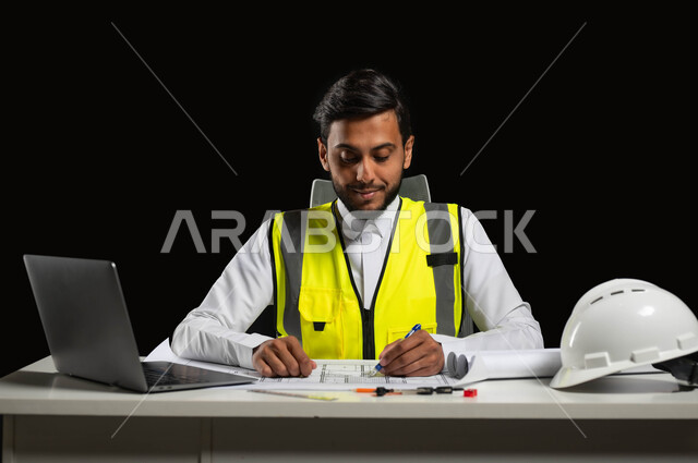 مراجعة الخطط الهندسية, دور الهندسة الإنشائية في تطور المجتمع السعودي, بورتريه مقرب لمهندس عربي خليجي سعودي يرتدي الثوب التقليدي والسترة الخاصة بالعمل يجلس على المكتب ويمسك بيده القلم على المخطط الهندسي ، خلفية سوداء، محتوى حساس، استخدام حساس