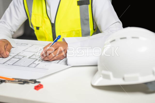 تدقيق المخططات والتصاميم المعمارية, بورتريه مقرب  لمهندس عربي خليجي سعودي يرتدي الثوب التقليدي و سترة الحماية يسجل على مخطط الإعمار, إدارة المشاريع الهندسية, مهن ووظائف سعودية, العمل في القطاع الهندسي, خلفية سوداء، محتوى حساس، استخدام حساس