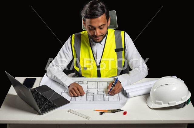 تدقيق ومراجعة الخطط الهندسية, مفهوم الهندسة والإنشاءات، بورتريه مقرب لمهندس عربي خليجي سعودي يرتدي الثوب التقليدي والسترة الخاصة بالعمل يجلس على المكتب ويمسك بيده القلم على المخطط الهندسي ، إكمال رسم مخطوطات البناء، خلفية سوداء، محتوى حساس، استخدام حساس