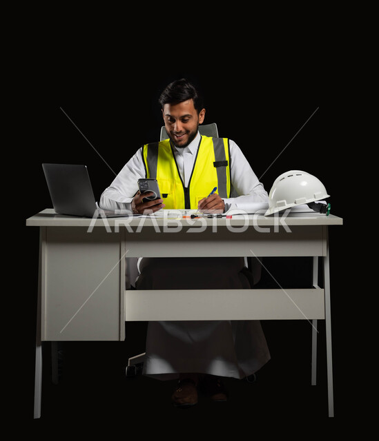 انجاز مهام العمل عبر الجهاز المحمول، مواكبة التطورات الحديثة, بورتريه مقرب لمهندس عربي خليجي سعودي يرتدي الثوب التقليدي مع سترة الحماية يقوم بالتعديل على مخطط الإعمار عن طريق بعض التطبيقات والمواقع, خلفية سوداء،الجسم بالطول الكامل، استخدام حساس، محتوى حساس