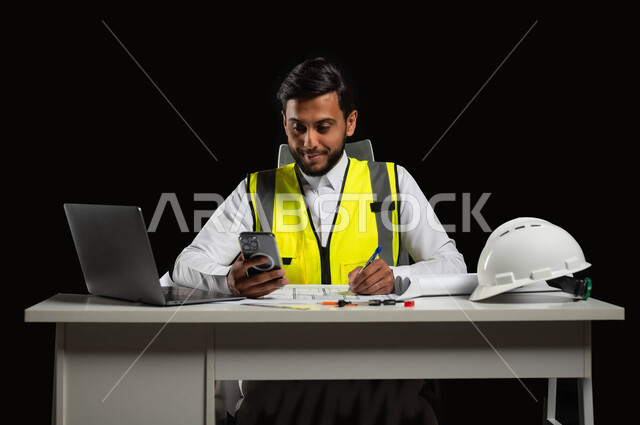 Developing and developing the engineering fields, keeping up with modern developments, a close-up portrait of a Saudi Gulf Arab engineer wearing the traditional dress with a protective jacket, amending the reconstruction plan through some applications and websites, black background, sensitive use, sensitive content