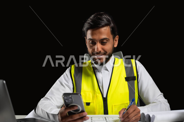 دمج التكنولوجيا والتقنية بالعمل,، مواكبة التطورات الحديثة, بورتريه مقرب لمهندس عربي خليجي سعودي يرتدي الثوب التقليدي مع سترة الحماية يقوم بالتعديل على مخطط الإعمار عن طريق بعض التطبيقات والمواقع, خلفية سوداء، استخدام حساس، محتوى حساس