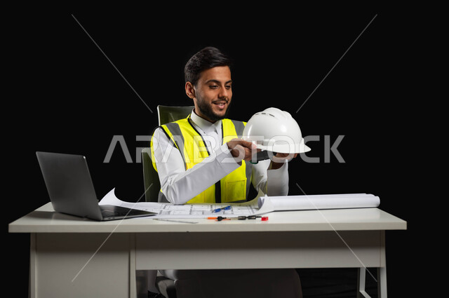 مفهوم الإنشاءات و الهندسة، النظر الى خوذه الحمايه البيضاء بايماءات السرور والفخر، بورتريه مقرب لرجل عربي خليجي سعودي يرتدي الثوب التقليدي مع السترة الخاصة بالعمل، مهن ووظائف سعودية, خلفية سوداء، محتوى حساس, استخدام حساس