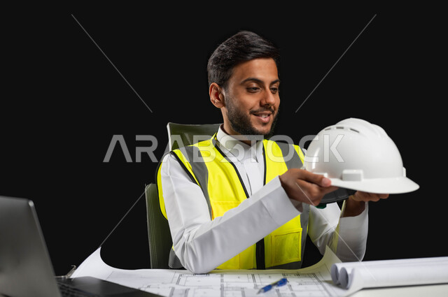 ادارة المشاريع الهندسية، النظر الى خوذه الحمايه البيضاء بايماءات السرور والفخر، بورتريه مقرب لرجل عربي خليجي سعودي يرتدي الثوب التقليدي مع السترة الخاصة بالعمل، مهن ووظائف سعودية, خلفية سوداء، محتوى حساس, استخدام حساس