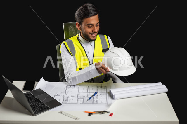 مهن ووظائف سعودية, النظر الى خوذه الحمايه البيضاء بايماءات السرور والفخر، بورتريه مقرب لرجل عربي خليجي سعودي يرتدي الثوب التقليدي مع السترة الخاصة بالعمل يجلس خلف المكتب ، العمل في المجال الهندسي،خلفية،سوداء محتوى حساس, استخدام حساس