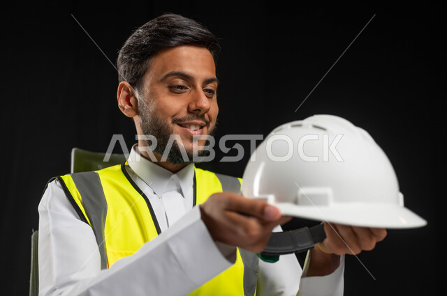 العمل في المجال الهندسي، النظر الى خوذه الحمايه البيضاء بايماءات السرور والفخر، بورتريه مقرب لرجل عربي خليجي سعودي يرتدي الثوب التقليدي مع السترة الخاصة بالعمل، مهن ووظائف سعودية, خلفية سوداء، محتوى حساس, استخدام حساس