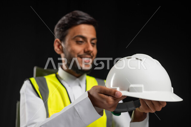 النظر الى خوذه الحمايه البيضاء بايماءات السرور والفخر، بورتريه مقرب لرجل عربي خليجي سعودي يرتدي الثوب التقليدي مع السترة الخاصة بالعمل، العمل في المجال الهندسي، مهن ووظائف سعودية, خلفية سوداء، محتوى حساس, استخدام حساس