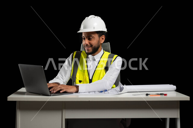 متابعة المشاريع الإنشائية, دراسة أساسيات المشروع عبر تطبيقات الحاسوب المحمول، التكنولوجيا والمجالات الهندسية,  بورتريه مقرب لمهندس عربي خليجي سعودي يرتدي الثوب التقليدي مع الخوذة وسترة الحماية يجلس خلف المكتب، خلفية سوداء، استخدام حساس، محتوى حساس