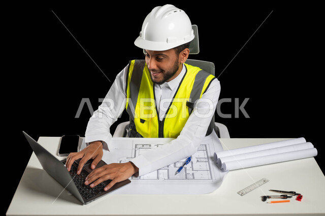 دراسة أساسيات المشروع عبر تطبيقات الحاسوب المحمول، متابعة المشاريع الإنشائية, التكنولوجيا والمجالات الهندسية,  بورتريه مقرب لمهندس عربي خليجي سعودي يرتدي الثوب التقليدي مع الخوذة وسترة الحماية يجلس خلف المكتب، خلفية سوداء، استخدام حساس، محتوى حساس