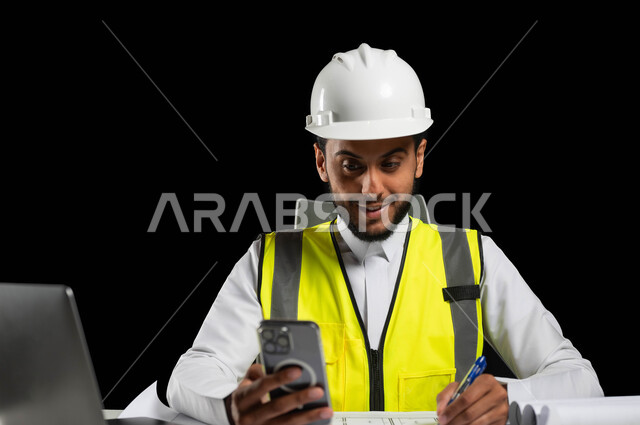 النظر الى شاشة الهاتف المحمول بايماءات السرور، تسجيل الملاحظات على اوراق العمل، دمج التقنية والتكنولوجيا في مجال الهندسة، بورتريه مقرب لمهندس عربي خليجي سعودي يرتدي الثوب التقليدي مع الخوذة وسترة الحماية يجلس خلف المكتب، خلفية سوداء، استخدام حساس، محتوى حساس