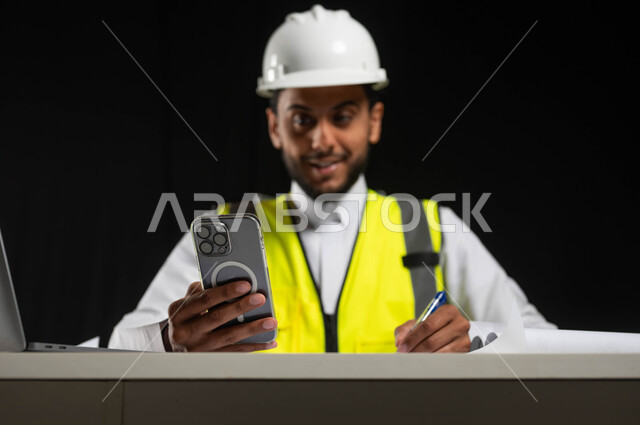 Using modern and advanced technologies, looking at the mobile phone screen with pleased gestures, recording notes on work papers, a close-up portrait of a Saudi Gulf Arab engineer wearing a traditional dress with a helmet and a protective jacket sitting behind the desk, black background, sensitive use, sensitive content