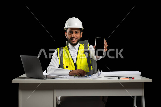 Integrating technology and technology in the field of engineering, looking at the camera with gestures of pleasure, displaying a blank black screen, close-up portrait of an Arab engineer, a Saudi Khalidi, wearing the traditional dress with a helmet and a protective vest, sitting behind the desk, black background, sensitive use, sensitive content