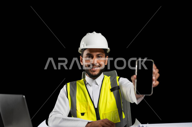 استخدام التقنيات الحديثة لتسهيل القيام بالمهام المطلوبة, دمج العمل مع التكنولوجيا، عرض شاشة جوال سوداء,بورتريه لمهندس عربي خليجي سعودي يرتدي الخوذة البيضاء وسترة الحماية الخاصة بالعمل, النظر للكاميرا بثقة مع إيماءلت الفرح والسعادة, خلفية سوداء