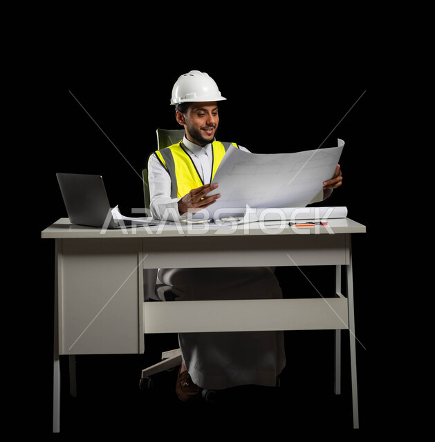 Studying the basics of the construction plant, work in the engineering sector, portrait of a Saudi Gulf Arab engineer wearing a protective vest and helmet holding construction plans in his hands, looking at the plan with great pride and happiness, reviewing and displaying project manuscripts completely, project management and follow-up, black background