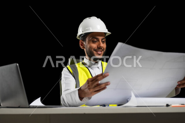 Reconstruction with the help of the nation’s people, work in the engineering sector, portrait of a Saudi Gulf Arab engineer wearing a protective vest and helmet holding reconstruction plans in his hands, looking at the plan with pride and great happiness, reviewing and displaying project manuscripts in full, project management and follow-up, black background