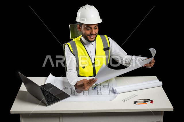 مفهوم الهندسة والانشاءات المعمارية, العمل القطاع الهندسي, بورتريه لمهندس عربي خليجي سعودي يرتدي سترة الحماية والخوذة يمسك مخططات الإعمار بيديه, النظر إلى المخطط بفخر وسعادة عارمة، مراجعة وعرض مخطوطات المشاريع بشكل كامل, إدارة ومتابعة المشروع, خلفية سوداء