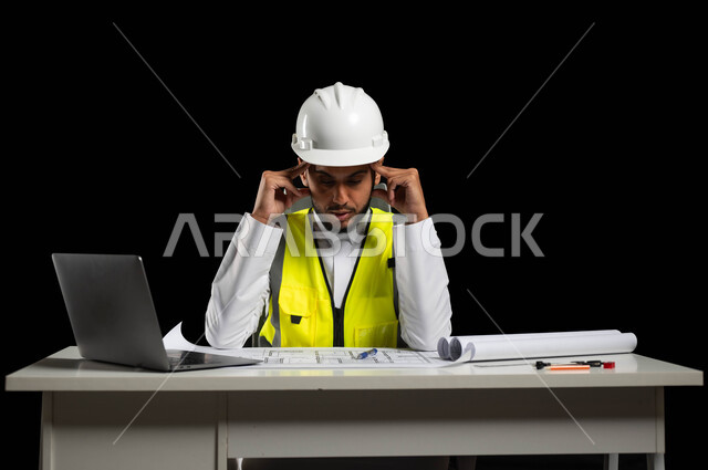 الإشراف على الأعمال وإدارتها في المكتب الهندسي, بورتريه لمهندس عربي خليجي سعودي يرتدي الخوذة البيضاء وسترة الحماية الخاصة بالعمل يجلس وراء المكتب ويمسك رأسه بيديه بإيماءات تدل على الشعور بالصداع, الحاجة إلى الراحة والتعافي، خلفية سوداء