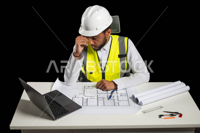إدارة ودراسة المشاريع الهندسية، متابعة الخطط والتعليمات, بورتريه لمهندس عربي خليجي سعودي يرتدي الخوذة البيضاء وسترة الحماية الخاصة بالعمل يجلس وراء المكتب ويضع يده على رأسه بإيماءات تدل على التعب والإرهاق, الجد والاجتهاد في العمل، خلفية سوداء