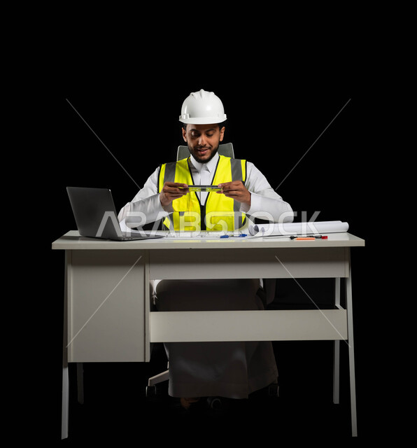 الاستمتاع في الأعمال الهندسية ومتابعة المشاريع الإنشائية, سترة خاصة بالعمل وخوذة بيضاء للحماية, بورتريه لمهندس انشاءات عربي خليجي سعودي يجلس وراء طاولته المكتبية يمسك بيده أدوات هندسية, الحاسوب المحمول والمخططات أمامه, النظر بإيماءات السعادة والفرح, خلفية سوداء
