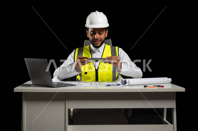 استثمار التكنولوجيا في المجال الهندسي, سترة خاصة بالعمل وخوذة بيضاء للحماية, بورتريه لمهندس انشاءات عربي خليجي سعودي يجلس وراء طاولته المكتبية يمسك بيده أدوات هندسية, عرض لمخططات البناء أمامه, النظر بإيماءات الفخر والسعادة, خلفية سوداء