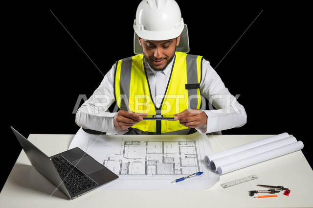 العمل في المكتب الهندسي, سترة خاصة بالعمل وخوذة بيضاء للحماية, بورتريه لمهندس انشاءات عربي خليجي سعودي يجلس وراء طاولته المكتبية يمسك بيده أدوات هندسية, عرض مخطط البناء بشكل كامل, النظر بإيماءات الفخر والسعادة, خلفية سوداء