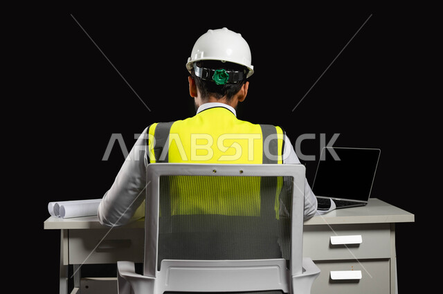 تطور المجال الهندسي في المملكة العربية السعودية بسواعد أبنائها, بورتريه من الخلف لمهندس معماري  عربي خليجي سعودي يرتدي الخوذة البيضاء وسترة العمل يجلس على الكرسي وأمامه المكتب, جهاز الحاسوب المحمول وبعض المخططات المعمارية على الطاولة الهندسية, خلفية سوداء