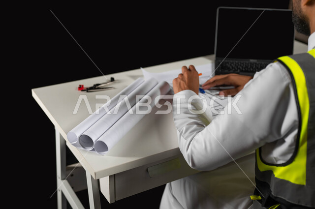بحث حيثيات المشروع وتدوين الملاحظات الهامة,تدقيق ومراجعة الخطط الهندسية, مفهوم الهندسة والإنشاءات المعمارية, بورتريه مقرب من الجانب لمهندس عربي خليجي سعودي يرتددي سترة الحماية الخاصة بالعمل ويقوم بكتابة التعديلات على أوراق مخطط البناء, خلفية سوداء