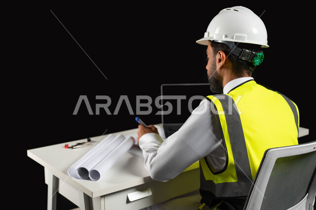 التعديل على خطط المشروع ومتابعة العمل, مفهوم الهندسة الإنشائية، بورتريه لمهندس عربي خليجي سعودي يرتدي الخوذة البيضاء والسترة الخاصة بالعمل يجلس على المكتب بيده القلم ويرسم المخطط الهندسي، الحاسوب المحمول والمخطوطات المعمارية على الطاولة المكتبية, خلفية سوداء