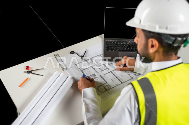إكمال رسم مخطوطات البناء, مفهوم الهندسة والإنشاءات المعمارية، بورتريه لمهندس عربي خليجي سعودي يرتدي الخوذة البيضاء والسترة الخاصة بالعمل يجلس على المكتب بيده القلم ويرسم المخطط الهندسي، الحاسوب المحمول والمخطوطات المعمارية على الطاولة المكتبية, خلفية سوداء