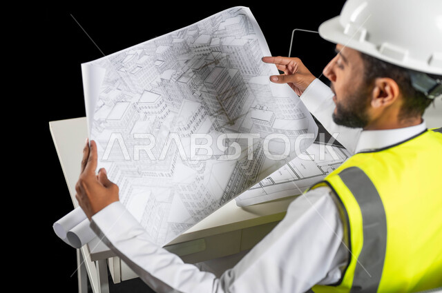 مراجعة الخطط الهندسية, دور الهندسة الإنشائية في تطور المجتمع السعودي, بورتريه مقرب لمهندس عربي خليجي سعودي يرتدي خوذة الحماية والسترة الخاصة بالعمل يجلس على المكتب ويمسك بيديه مخطط البناء, العرض الكامل لمخطط المشروع, الإشراف على سير العمل, خلفية سوداء