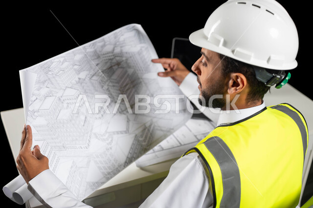 دور الهندسة الإنشائية في تطور المجتمع السعودي, متابعة سير العمل, عرض مخطط المشروع بشكل كامل, بورتريه لمهندس عربي خليجي سعودي يرتدي خوذة الحماية والسترة الخاصة يجلس على المكتب ويمسك بيديه المخطط, إدارة ومتابعة عملية البناء، خلفية سوداء
