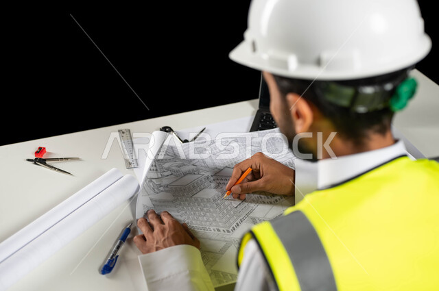 تدقيق ومراجعة الخطط الهندسية, مفهوم الهندسة والإنشاءات المعمارية، بورتريه مقرب من الخلف لمهندس عربي خليجي سعودي يرتدي الخوذة البيضاء والسترة الخاصة بالعمل يجلس على المكتب ويمسك بيده القلم ويرسم المخطط الهندسي ، إكمال رسم مخطوطات البناء، خلفية سوداء