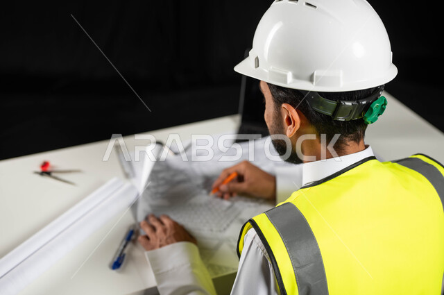 متابعة استراتيجات المشروع, مفهوم الهندسة والإنشاءات المعمارية، بورتريه لمهندس عربي خليجي سعودي يرتدي الخوذة البيضاء والسترة الخاصة بالعمل يجلس على المكتب ويمسك بيده القلم ويرسم المخطط الهندسي ، إكمال رسم مخطوطات البناء، تدقيق المشاريع ومراجعتها، خلفية سوداء