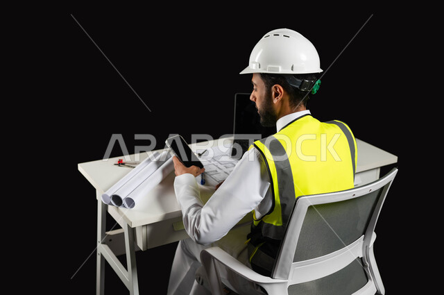 تصفح التطبيقات والمواقع الهندسية, بورتريه لمهندس عربي خليجي سعودي يرتدي سترة الحماية و الخوذة  البيضاء يجلس وراء المكتب وينظر إلى شاشة الجوال, هندسة الأبنية و الإنشاءات، مخططات البناء  والحاسوب المحمول على الطاولة المكتبية, خلفية سوداء
